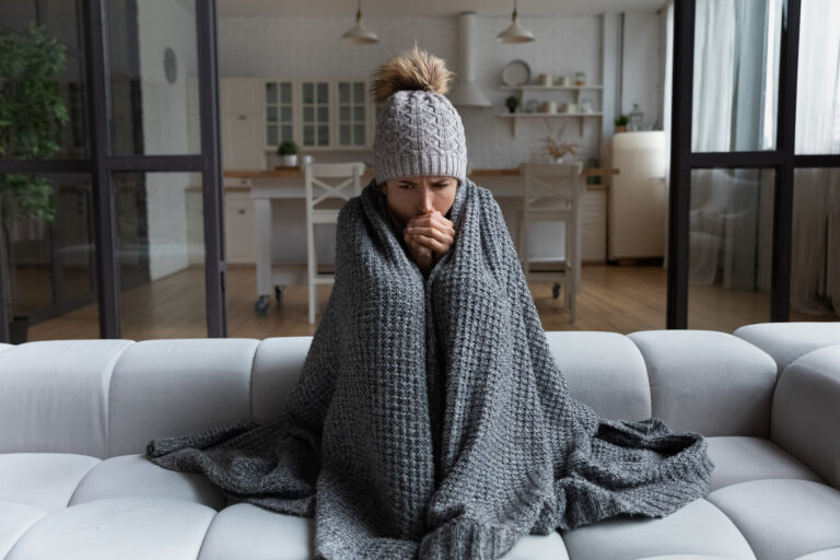 Cold woman on couch with blanket and hat, blowing on her hands